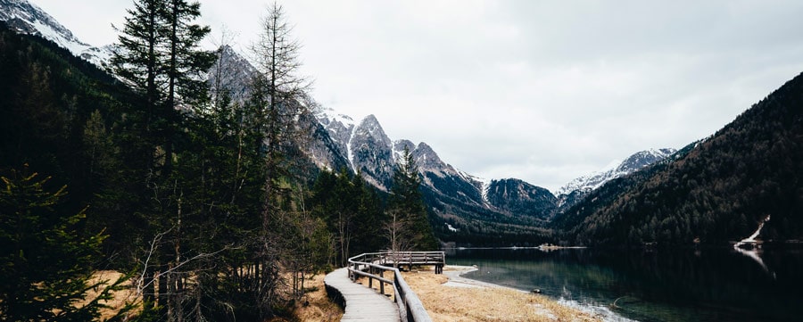 Wooden path next to a lake leading towards mountains with snowy peaks.